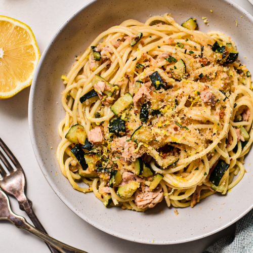 Zucchini tuna pasta with lemon served in a white shallow bowl next to forks and a lemon half.