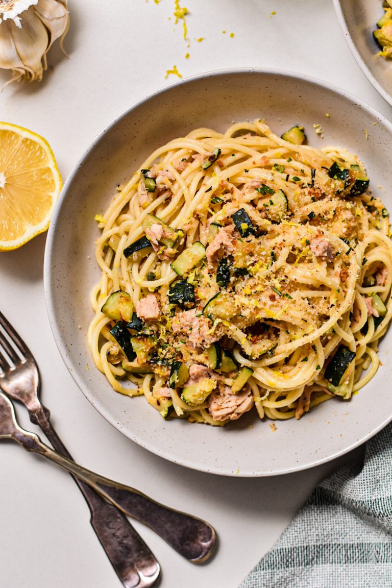 Zucchini tuna pasta with lemon served in a white shallow bowl next to forks and a lemon half.