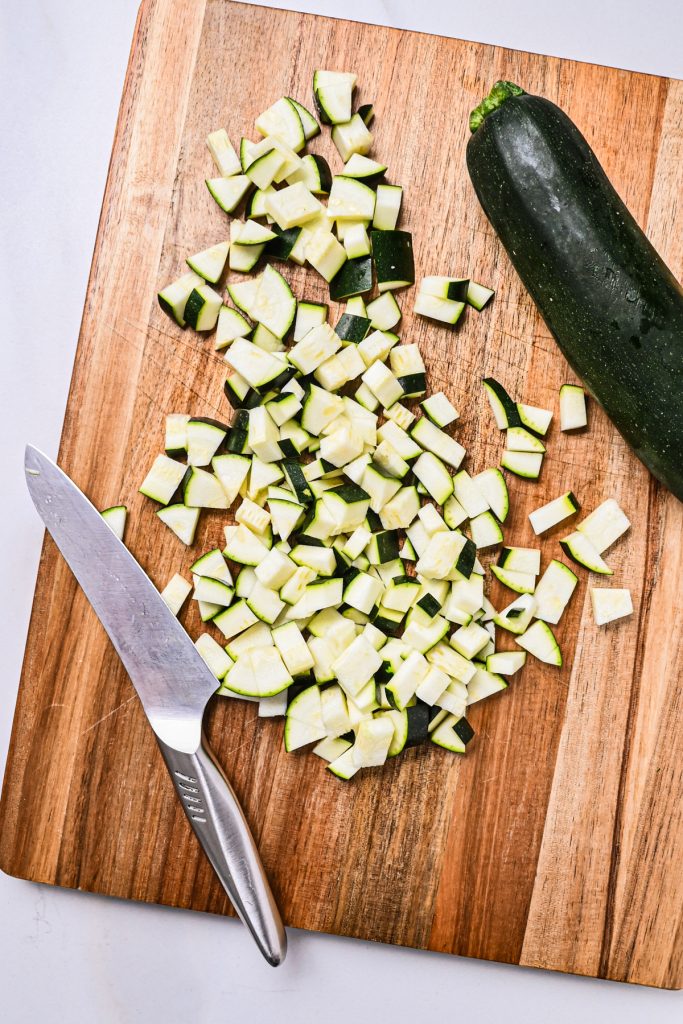Diced zucchini on a wooden cutting board next to a knife.