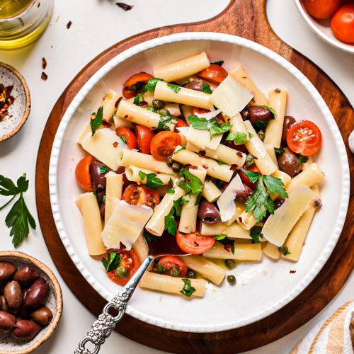 Puttanesca pasta salad in a serving bowl on a round wooden board.