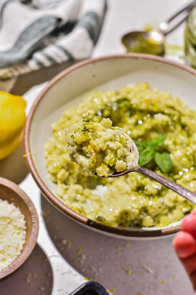 Hand holding a spoon with a bite of the creamy pesto pastina on it and more in a bowl in the background.