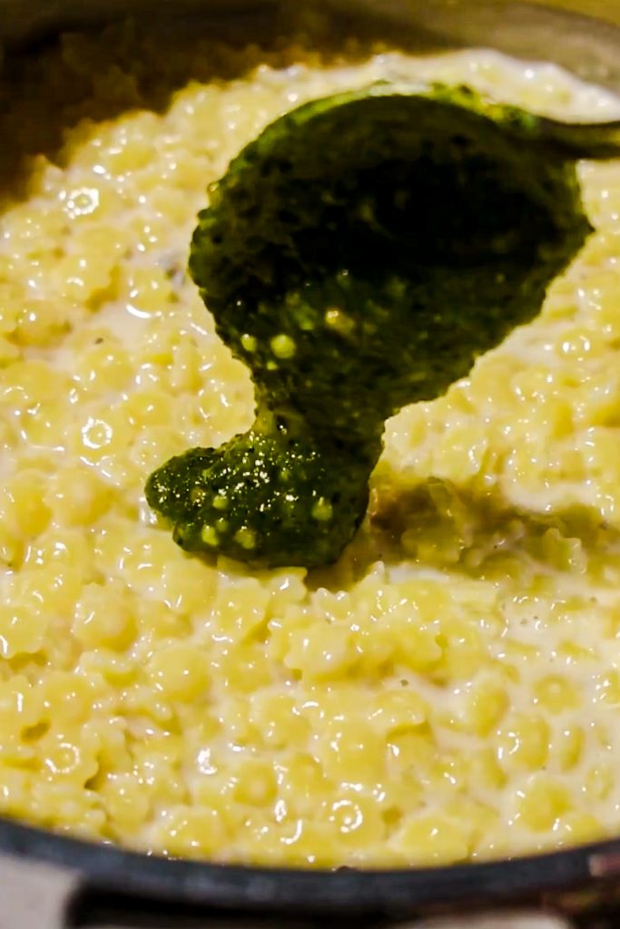 Using a spoon to add the pesto into the creamy pastina in a pot.