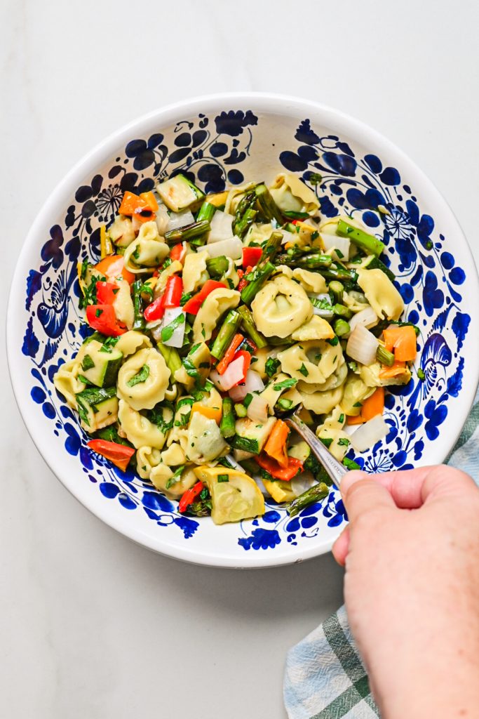 Hand stirring the grilled veggie tortellini pasta salad in a bowl.